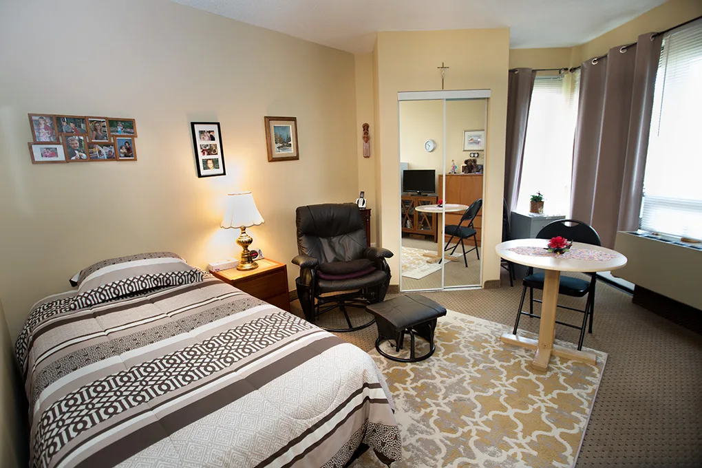 A bright resident room with a bed, reading chair, table for two and large curtained windows