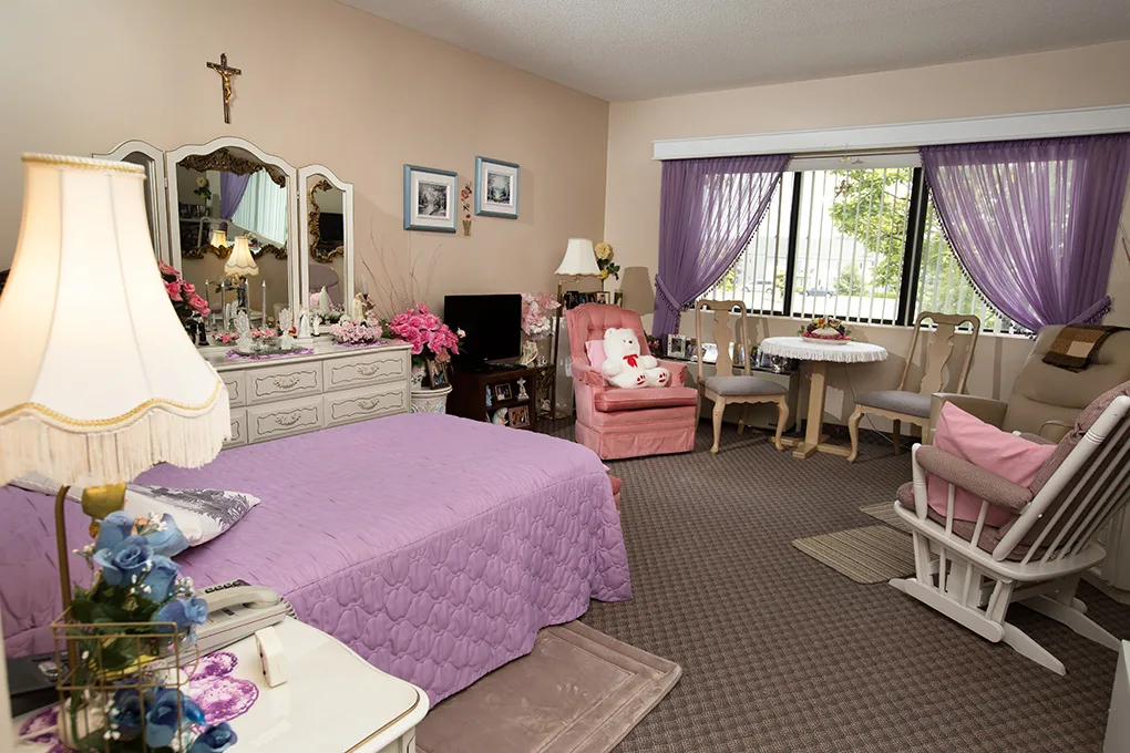 The Single Suite with Village View — a resident's suite furnished with dresser, sitting chairs and a wide window